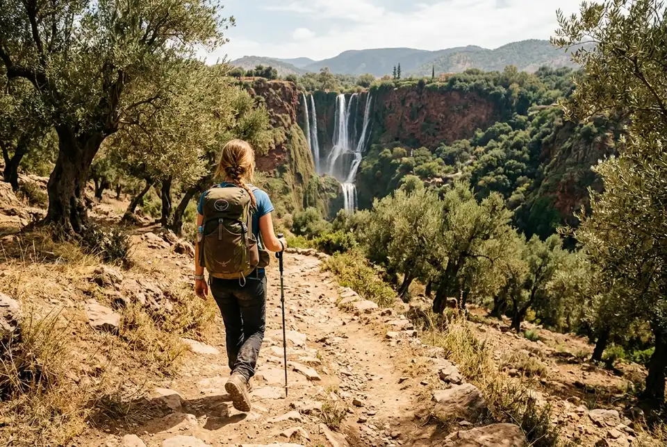 Solo traveler with a backpack and hiking shoes walking an uneven olive-grove trail toward Ouzoud Waterfalls, no guide in sight, midday light